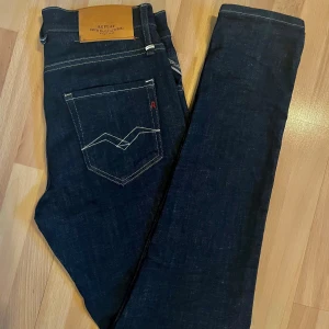 Replay Anbass mörkblå jeans - Snygga mörkblå Replay Anbass jeans med klassisk femficksdesign och kontraststickningar. Modellen har raka ben och normal passform, med läderpatch bak i midjan och diskret Replay-logga vid fickan. Perfekta för dig som gillar stilrena och tidlösa jeans. Använda en gång