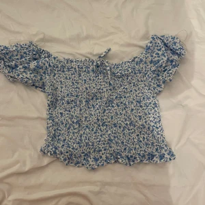 Blommig offshoulder topp från FB Sister - Supersöt offshoulder topp från FB Sister i storlek L. Toppen är vit med blått blommönster, har smockad kropp och volangkant nedtill. Puffiga korta ärmar och en liten knytning framtill ger en romantisk vibe. Perfekt för varma dagar och chill häng.