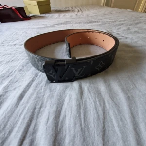 Louis Vuitton svart skinnbälte - Säljer ett Louis Vuitton bälte i svart skinn med klassiskt monogrammönster och en stilren LV-spänne i svart metall. Insidan är ljusbrun och har präglad logga. Bältet är brett och har flera hål för justering. Perfekt för dig som gillar exklusiva accessoarer.