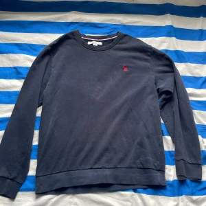 Mörkblå sweatshirt från U.S. Polo Assn. - Snygg mörkblå sweatshirt från U.S. Polo Assn. med klassisk rund hals och diskret röd logobroderi på bröstet. Tröjan har långa ärmar och ribbade muddar vid ärmslut och nederkant. Perfekt för chill dagar och enkel att matcha med jeans eller joggers. Smått urtvättad på ett ställe de bild 4. Passar både M och L