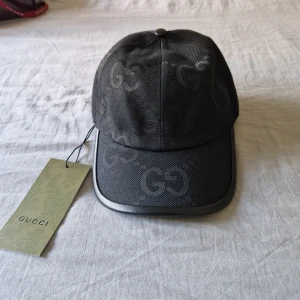 Svart keps från Gucci med GG-logga - Snygg svart keps från Gucci med GG-monogram över hela kepsen. Kepsen har böjd skärm med kant i svart och justerbar rem bak i skinn. Materialet är robust och har en lyxig känsla, perfekt för dig som vill ha en statement-accessoar.