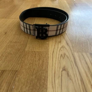 Burberry rutig bälte med B-spänne - Säljer ett snyggt Burberry bälte med klassiskt rutigt mönster i beige, brun och svart. Bältet har ett stilrent svart metallspänne med Burberrys ikoniska B-logga. Materialet är skinn med präglad struktur och exklusiv känsla. Perfekt för dig som vill ha en statement-accessoar.