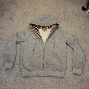 Grå Burberry hoodie - Säljer en grå hoodie från Burberry med klassiskt rutigt mönster i huvan och insidan. Tröjan har dragkedja, snörning vid huvan och broderad Burberry-logga på bröstet. Mjuk bomullskvalitet och två fickor framtill. Perfekt för dig som gillar stilren design med lyxig touch. Använd tre gånger