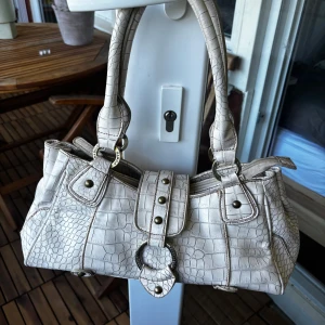 Vit handväska med krokopräglat mönster - Snygg vit handväska från Babila med krokopräglat mönster och coola metalldetaljer. Väskan har två rejäla handtag, dekorativa nitar och ringar med Babila-logga. Flera fack med dragkedja och en unik form som sticker ut. Perfekt för dig som gillar statement-accessoarer.Bonus den är i äkta krokodil skinn.