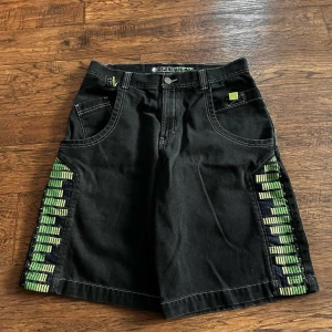 Svarta EQ shorts med gröna detaljer - Unika svarta shorts från EQ med gröna och gula grafiska detaljer längs sidorna och broderad logga på bakfickan. Modellen har breda ben, kontrastsömmar och flera fickor. Perfekt för dig som gillar streetwear och vill sticka ut med cool design.