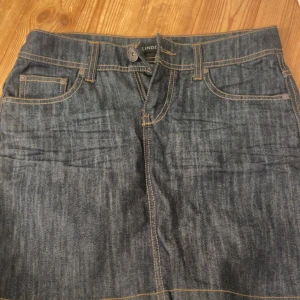 Mörk jeanskjol från Lindex - Snygg mörkblå jeanskjol från Lindex med klassiska fickor fram och bak, samt coola detaljer på bakfickorna. Kjolen har synliga sömmar i kontrastfärg och bälteshällor. Perfekt för dig som gillar en enkel men trendig look.