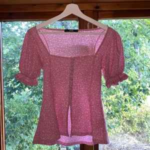 Rosa blommig blus från BikBok - Supersöt rosa blus från BikBok med små vita blommor, puffiga korta ärmar och knappar framtill. Blusen har en fyrkantig urringning och volangdetaljer vid ärmsluten:)