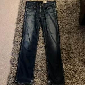 Wrangler Ace mörkblå jeans - Mörkblå jeans från Wrangler Ace med klassisk femficksdesign och snygga slitningar. Jeansen har raka ben och normal passform, med detaljer som kopparfärgade nitar och läderpatch bak. Perfekta för dig som gillar en tidlös och avslappnad look.