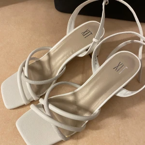 Vita sandaler med klack från XIT - Snygga vita sandaler från XIT med fyrkantig tå och trendiga remmar som korsar över foten. Skorna har en stabil blockklack och justerbar rem runt ankeln. Tillverkade i syntetmaterial med en stilren och modern look.
