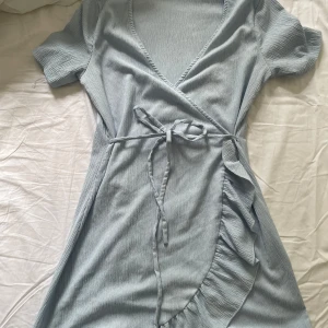 Ljusblå klänning - Supersöt ljusblå omlottklänning från Vero Moda i storlek S. Klänningen har korta ärmar, v-ringning och knytband i midjan. Volangdetaljer längs kanten ger en extra touch. Använd max 2 gånger, och kommer inte till användning längre därför jag säljer🥰
