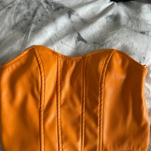 Orange korsett-topp i skinnimitation - Säljer en snygg orange korsett-topp i skinnimitation med markerade sömmar och lätt böjd sweetheart-urringning. Toppen är axelbandslös och har dragkedja i sidan för enkel påklädning. Perfekt för dig som vill sticka ut med färg och form!