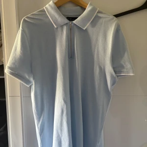 Ljusblå polo t-shirt med zip från S.C.W - Fräsch ljusblå polo t-shirt från S.C.W med vit kant på kragen och ärmslut. T-shirten har korta ärmar och en snygg dragkedja framtill. Perfekt för dig som gillar stilren och enkel design. Materialet är mjuk bomull som känns skönt mot huden.