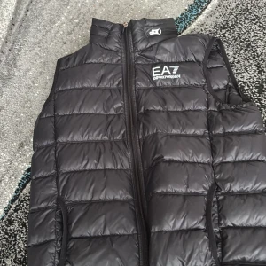 Svart dunväst EA7 Emporio Armani - Snygg svart dunväst från EA7 Emporio Armani med quiltad design och dragkedja framtill. Västen har hög krage, två fickor och avtagbar huva med logga. Perfekt för lager-på-lager och streetstyle.