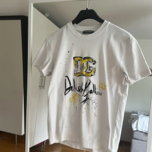 Vit Dolce & Gabbana t-shirt med print - Snygg vit t-shirt från Dolce & Gabbana med coolt grafiskt print i svart och gult framtill. Rund hals och klassisk passform. Materialet är mjuk bomull som känns skönt mot huden. Perfekt för dig som gillar streetstyle och exklusiva märken som även passar perfekt till fester! 