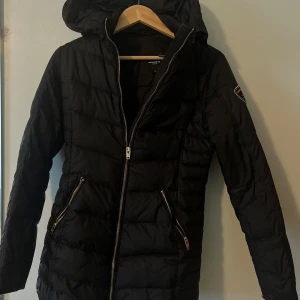 Svart pufferjacka Rockandblue 34 - Säljer en svart pufferjacka från Rockandblue i storlek 34. Pälskrage medföljer. Jackan har huva, två snedställda fickor med dragkedja och Rockandblue-märke på ärmen. Perfekt för kyliga dagar och har en snygg quiltad look med långa ärmar och hel dragkedja framtill.