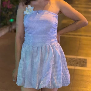 Ljusblå axelbandslös kortklänning - Söt ljusblå och vit axelbandslös kortklänning med rutigt mönster. Klänningen har en voluminös kjol och en dekorativ blomma vid bröstet. Perfekt för sommarens varma dagar och har en ungdomlig, fräsch vibe.