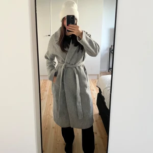 Grå kappa från Mango - Snygg grå kappa från Mango med knytbälte i midjan och fickor på sidorna. Det är en lång modell och har ett mjukt och mysigt material, perfekt nu inför hösten.🍁