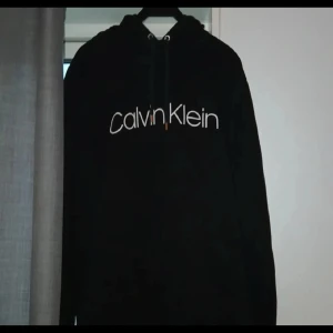 Svart Calvin Klein hoodie med logga - Svart hoodie från Calvin Klein med stor vit logga över bröstet. Klassisk modell med huva och snörning samt känguruficka framtill. Mjuk och skön, perfekt för chill dagar. Snygg och enkel att matcha med jeans eller joggers.