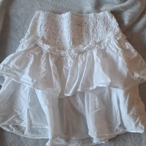 Vit volangkjol från BikBok - Supersöt vit kortkjol från BikBok med bred smock i midjan och flera lager volanger. Kjolen är gjord i tunn bomull och har en luftig känsla, perfekt för varma dagar. Enkel att matcha med crop tops eller stickade tröjor.