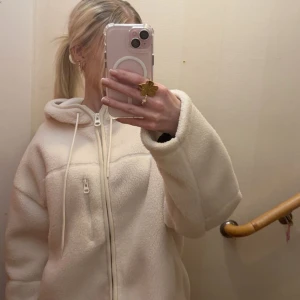 Vit teddyjacka med huva från FB Sister - Mysig vit teddyjacka från FB Sister i storlek S. Jackan har huva med snörning, hel dragkedja framtill och en extra ficka med dragkedja på bröstet. Tillverkad i mjuk fleece som ger en chill och avslappnad vibe. Perfekt för kalla dagar och enkel att styla. 🤍🤍