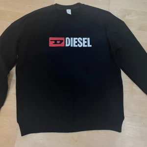 DIESEL TRÖJA - Svart sweatshirt från Diesel med stort vitt och rött logotryck på bröstet. Klassisk rund halsringning och långa ärmar. Perfekt för dig som gillar streetwear och vill ha en enkel men snygg tröja med tydlig branding.