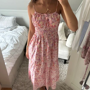 Blommig långklänning med smala axelband - Säljer en superfin långklänning med smala axelband och blommigt mönster i rosa, lila och rött. Klänningen har smock i midjan och en söt knytning vid halsen. Perfekt för varma dagar och har en luftig, flowy passform. Materialet känns lätt och bekvämt mot huden. Funkar även som kjol. 
