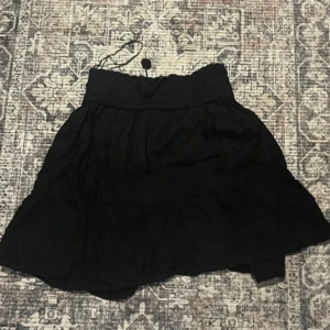 Svart kortkjol med volang - Säljer en svart kortkjol med bred, ribbad midja och volangdetaljer. Kjolen har en flowy och luftig passform som ger en skön vibe. Perfekt att matcha med en cool topp eller hoodie. Midjan har snörning för extra stilpoäng.