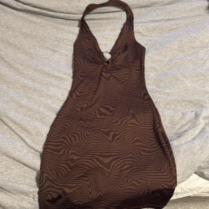 Brun zebramönstrad halterneckklänning - Säljer en snygg brun halterneckklänning från PLT i storlek XXS. Klänningen har ett coolt zebramönster och en ringdetalj framtill vid halsen. Den är figurnära och kort i modellen, perfekt för dig som gillar trendiga och unika plagg.