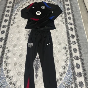 Svart Nike FC Barcelona half zip tracksuit.  - Svart tränings tracksuit från Nike x FC Barcelona med klubbmärke, Spotify-logga och half zip. Snygga detaljer i rött och blått på ärmarna. Tillverkad i 100% polyester, perfekt för fotbollsträning eller chill. Den har bara använts ett par gånger. PRIS KAN DISKUTERAS!