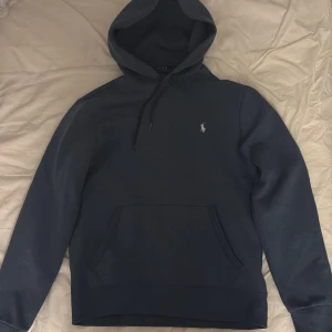 Ralph Lauren hoodie - Säljer åt min bror. Snygg marinblå hoodie från Polo Ralph Lauren med klassisk broderad logga på bröstet. Tröjan har huva med snörning och en stor magficka framtill. Storlek S. Mycket bra skick.