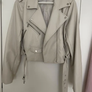 Beige skinnjacka från Stradivarius L - Snygg beige skinnjacka från Stradivarius i storlek L. Jackan har klassisk bikerstil med dragkedjor, knappar och bälte i midjan. Croppad modell med långa ärmar och coola detaljer som ger en edgy vibe till din outfit. Notera att jackan lyckats i tvätten fått en grå fläck i bakdelen av jackan därför säljs den så billigt, annars är den i väldigt gott skick.