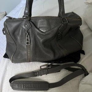 Grå väska från Zadig & Voltaire - Snygg grå handväska från Zadig & Voltaire med grov dragkedja och coola metalldetaljer. Väskan har både handtag och avtagbar axelrem med logga. Slitage är synligt💓