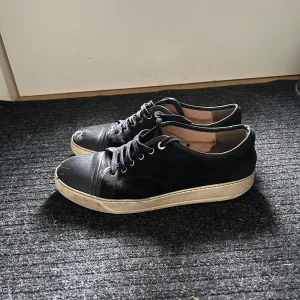 Svarta sneakers från Lanvin - Svarta sneakers från Lanvin med rund tå och platt vit sula. Ovandelen är i mocka med detaljer i skinn på tån och snörning framtill. Insidan är beige och har Lanvin-logga. Snygg och stilren modell som passar till jeans eller chinos.