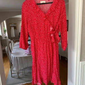 Röd omlottklänning från Holly & White - Snygg röd omlottklänning med vitt prickigt mönster från Holly & White. Klänningen har v-ringning, trekvartsärm och volangdetaljer nertill. Knyt i midjan för en fin siluett. Perfekt för dig som gillar färg och mönster!