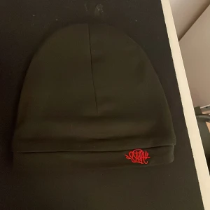 Svart mössa med röd broderad logga - Svart mössa från syna world med stilren design och uppvikt kant. På kanten finns en snyggt broderad logga i rött som ger en cool kontrast. Mössan är gjord i mjukt syntetmaterial och passar perfekt för dig som gillar streetwear.