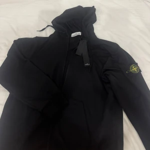 Svart hoodiejacka från Stone Island - Svart hoodiejacka från Stone Island med klassisk patch på ärmen. Jackan har dragkedja framtill, huva med snörning och ribbade muddar. Tillverkad i mjuk bomull för en skön och avslappnad stil. Perfekt för dig som gillar streetwear och vill ha något stilrent. Köpt av en annan säljare så ej säker på om den är äkta!