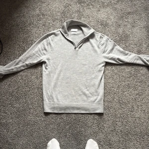 Grå merinoulltröja half zip These Glory Days - Ljusgrå långärmad tröja från These Glory Days i mjuk merinoull. Tröjan har en stilren half zip-dragkedja vid kragen och är stickad för en clean look. Perfekt för lager-på-lager och chill vardagsstil. Finns tyvärr två hål men ej något som syns vid användning. Storlek M men sitter mer som S