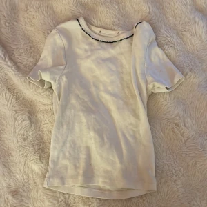 Vit croppad topp med svart kant - Säljer en vit croppad topp med korta ärmar och svart vågig kant runt halsen. T-shirten har en slim passform och är tillverkad i mjuk bomull. Perfekt för dig som gillar en clean och enkel stil med en liten twist.