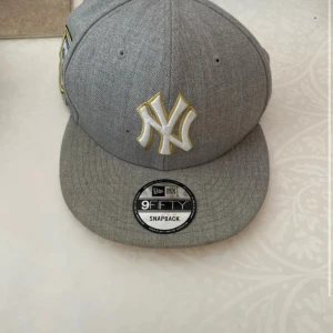 Grå New Era 9Fifty Snapback keps - Snygg grå snapback keps från New Era med klassiskt New York Yankees broderat i vitt och guld på framsidan. Modellen är 9Fifty med justerbar baksida och platt skärm. Perfekt för dig som vill ha en clean och sportig stil.