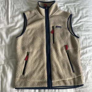 Beige fleeceväst från Patagonia - Säljer en beige fleeceväst från Patagonia i storlek S. Västen har marinblå detaljer, hel dragkedja framtill och tre fickor med röda zip-pullers. Perfekt för lager-på-lager och outdoor. Mjuk och fluffig känsla med hög krage. Säljer den så jag har vuxit ur den. Kvitto följer med.