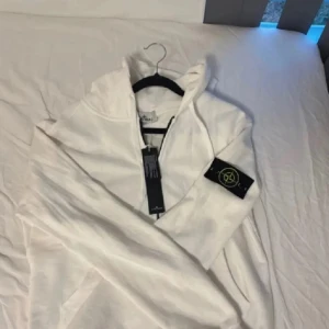 Vit kofta från Stone Island - Vit kofta från Stone Island med klassisk logga på ärmen. Tröjan har huva, hel dragkedja framtill och är tillverkad i mjuk bomull.🫶🏻