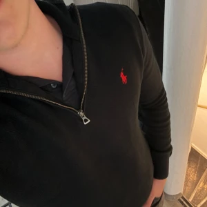 Svart half zip tröja Polo Ralph Lauren - Svart långärmad tröja från Polo Ralph Lauren i mjuk pima bomull. Klassisk half zip-dragkedja och röd broderad logga på bröstet. Snygg ribbad mudd vid ärmslut och nederkant. Perfekt för en clean och stilren look.
