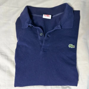 Marinblå pikétröja från Lacoste - Säljer en klassisk marinblå pikétröja från Lacoste. Piken har korta ärmar, krage och knappar framtill. Perfekt för en clean och sportig stil. Storlek M. 