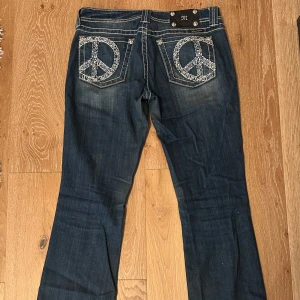 Miss Me bootcut jeans med peace-ryggfickor - Mörkblå bootcut jeans från Miss Me med kontrastsömmar och stora broderade peace-symboler på bakfickorna. Snygga detaljer med bling och vita dekorsömmar. Klassisk femficksmodell i jeansmaterial, perfekt för dig som gillar statement-plagg.