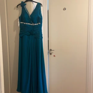 Turkos långklänning med strass  - Säljer en turkos långklänning från med v-ringad omlott upptill, strassdetaljer längs axel och midja samt dekorativa blommor på ena axeln. Klänningen har plisserad överdel och är ärmlös, perfekt för bal eller fest. Materialet känns lätt och elegant.