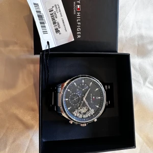 Blå och silver klocka Tommy Hilfiger - Snygg herrklocka från Tommy Hilfiger, helt ny! med blå urtavla och detaljer i silver. Klockan har en robust länkarmband i metall och flera små urtavlor för datum och veckodag. Stilren design med Hilfiger-logga och synliga mekaniska detaljer på sidan av urtavlan.