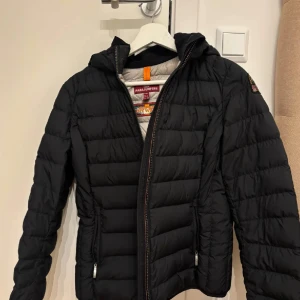 Svart dunjacka från Parajumpers Y-L - Säljer en svart, superlätt dunjacka från Parajumpers med huva och dragkedja. Jackan har quiltad design, två fickor med dragkedja och klassisk PJS-patch på ärmen. Perfekt för dig som vill ha en stilren och funktionell jacka med snygg passform.