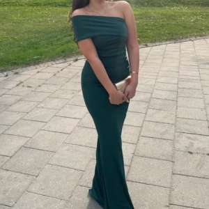 Mörkgrön offshoulder långklänning - Supersnygg mörkgrön långklänning med offshoulder och figurnära passform. Klänningen har en elegant, enkel design som framhäver siluetten och är perfekt för speciella tillfällen. Materialet känns mjukt och följsamt mot kroppen.