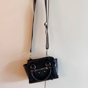 Svart croco axelväska med dragkedja - Snygg svart axelväska i croco-mönstrat skinnimitation med både handtag och justerbar axelrem. Väskan har silvriga metalldetaljer och en dragkedja framtill.. Perfekt storlek för det viktigaste. 
