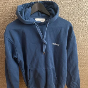 Les Deux hoodie - Snygg blå hoodie från Les Deux med broderad logga på bröstet. Tröjan har huva med snörning och en stor magficka. Perfekt för dig som gillar en enkel och stilren look. Mjuk bomullskvalitet och långärmad modell.